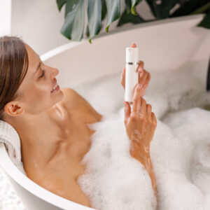 Joyful young woman holding body care product while bathing in bathtub with bubble foam