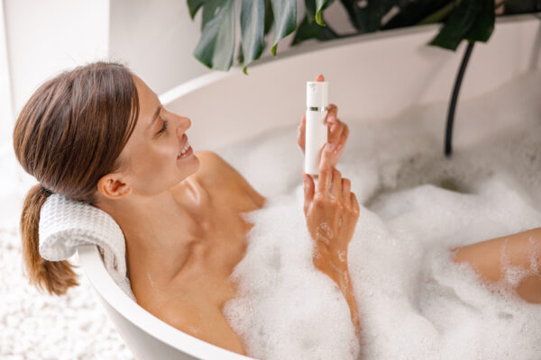 Joyful young woman holding body care product while bathing in bathtub with bubble foam