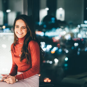 Portrait of young worker using phone, looking at the camera and smile at her workplace in evening.