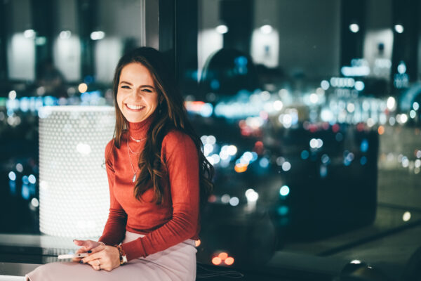 Portrait of young worker using phone, looking at the camera and smile at her workplace in evening.